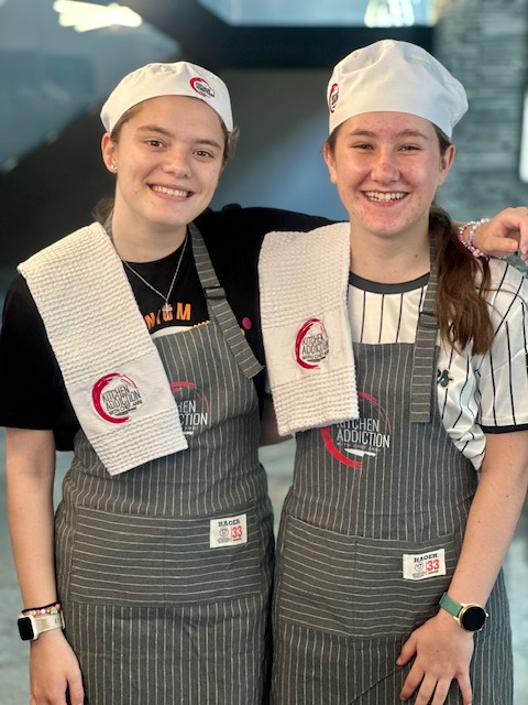 Two graduates smiling in their Kitchen Addiction aprons and chef hats