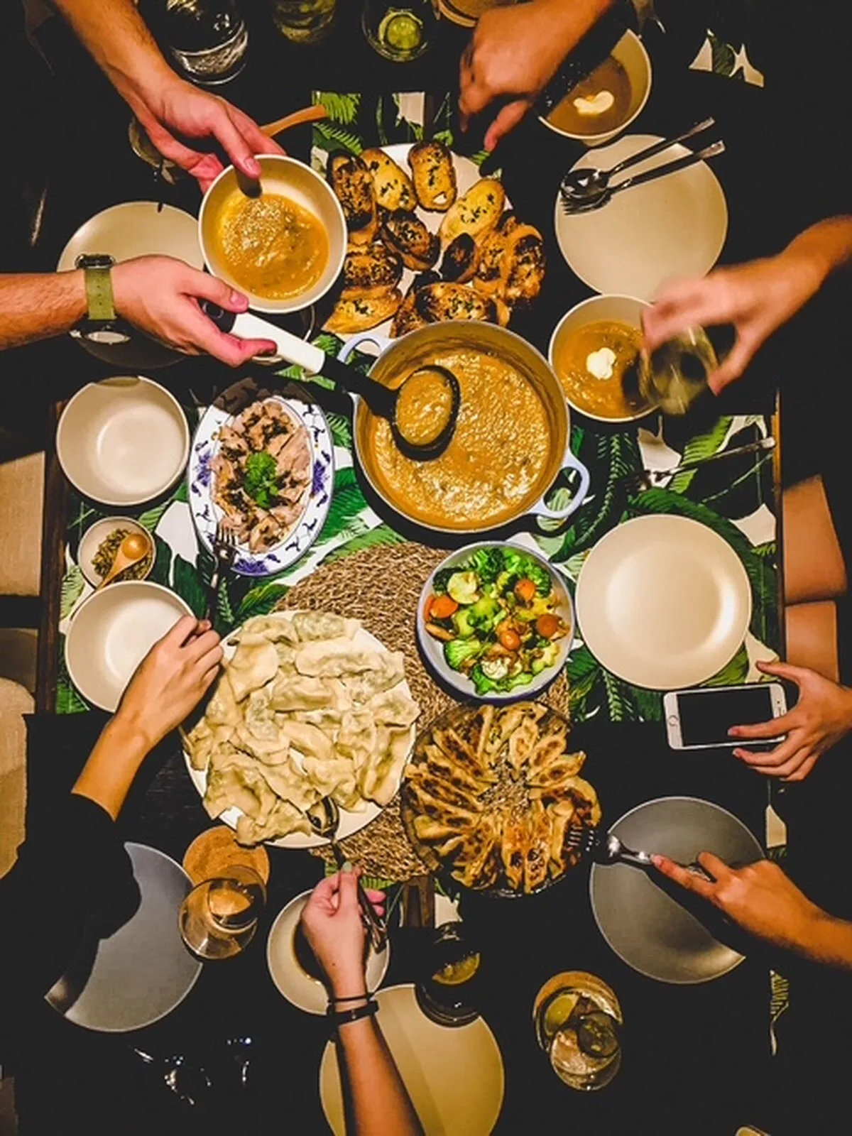 A Spanish table crowded with shared dishes — meatballs, pinchos, patatas bravas and dessert — as hands reach in to serve.