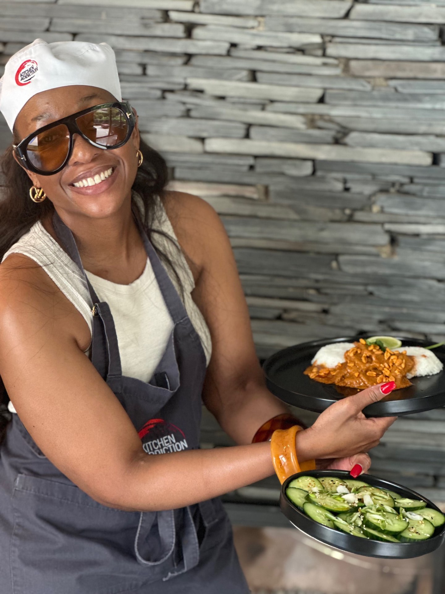 A guest wearing sunglasses and an apron holding two finished plates of curry with rice and a cucumber salad